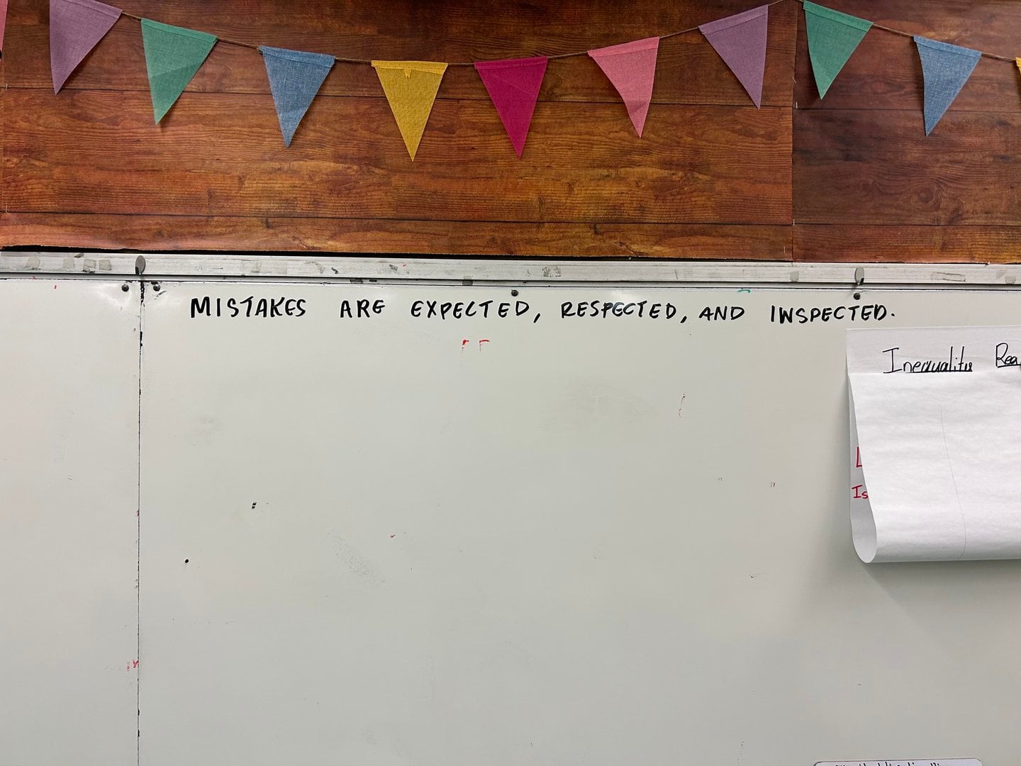 A whiteboard in a classroom that says "Mistakes are expected, respected, and inspected." There is a colorful triangle banner hanging above it.