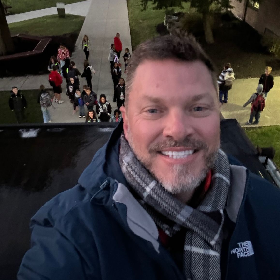 Adult standing outside on a roof, looking at camera. Behind adult below is a group of people