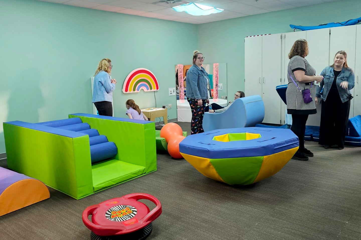 Several people sit and stand in a room full of sensory learning objects with blue walls in the background.