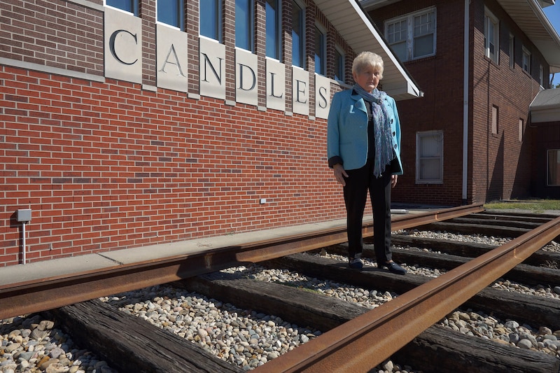 A woman with short white hair and wearing a bright blue jacket stands outside on a railroad track in front of a red brick building with the word "Candles" engraved on it.