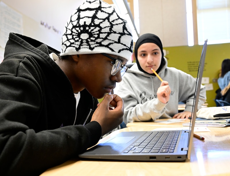 Two young students sit at a desk looking at a laptop in a classroom.