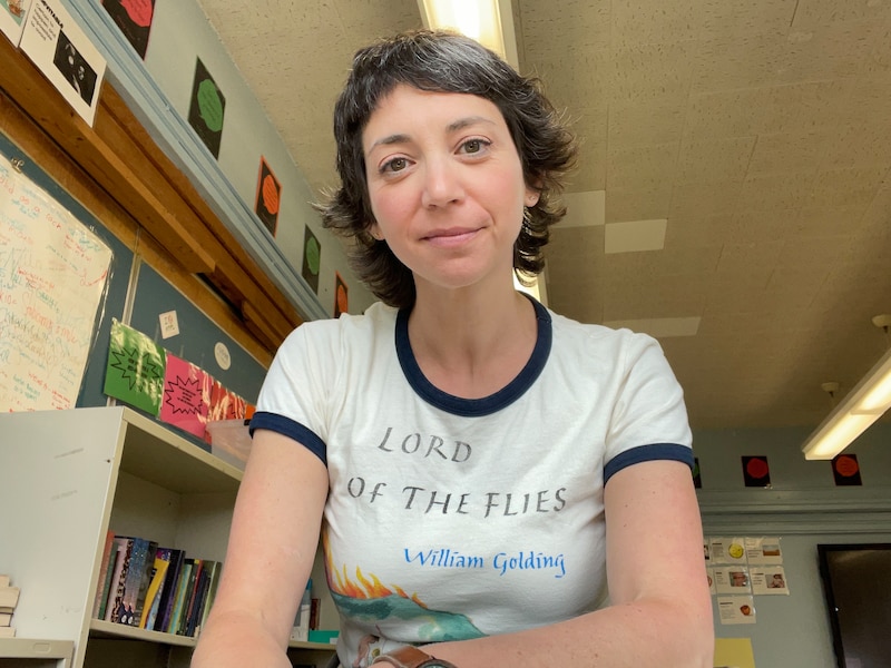 A woman with short dark hair and wearing a white t-shirt poses for a portrait in a classroom.