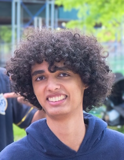 A high school student with short, dark and curly hair poses for a portrait outside with a tree and other people in the background.