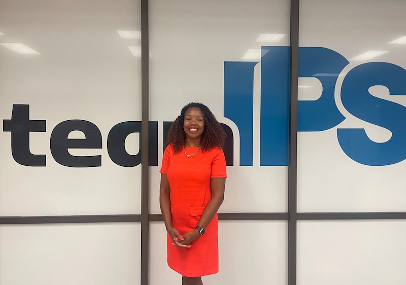 A woman in a vibrant orange outfit stands in front of a wall displaying the words "Team IPS."