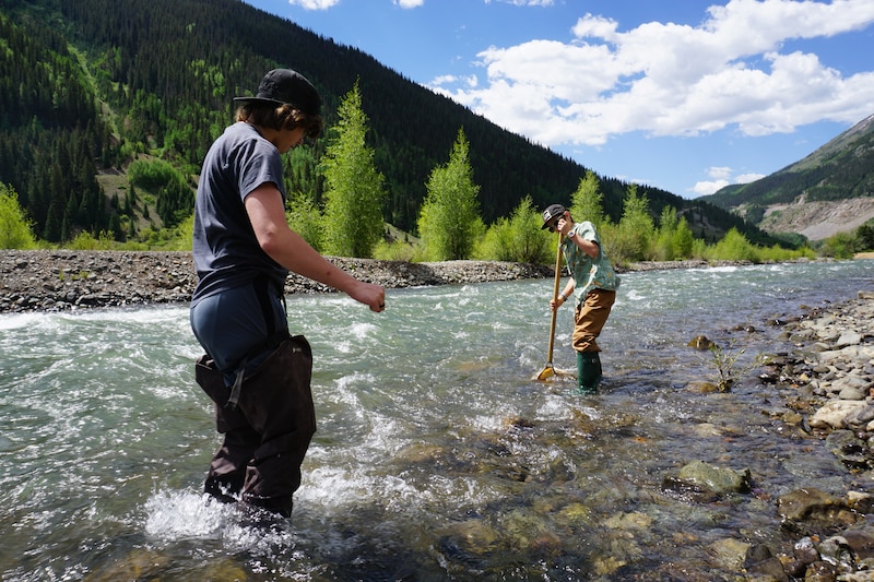 Two teenage boys in waders stand in a mountain stream.